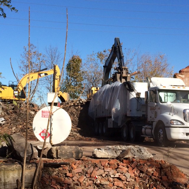Excavator loading demolition debris into a dump truck at a construction site with scattered brick rubble in the foreground.