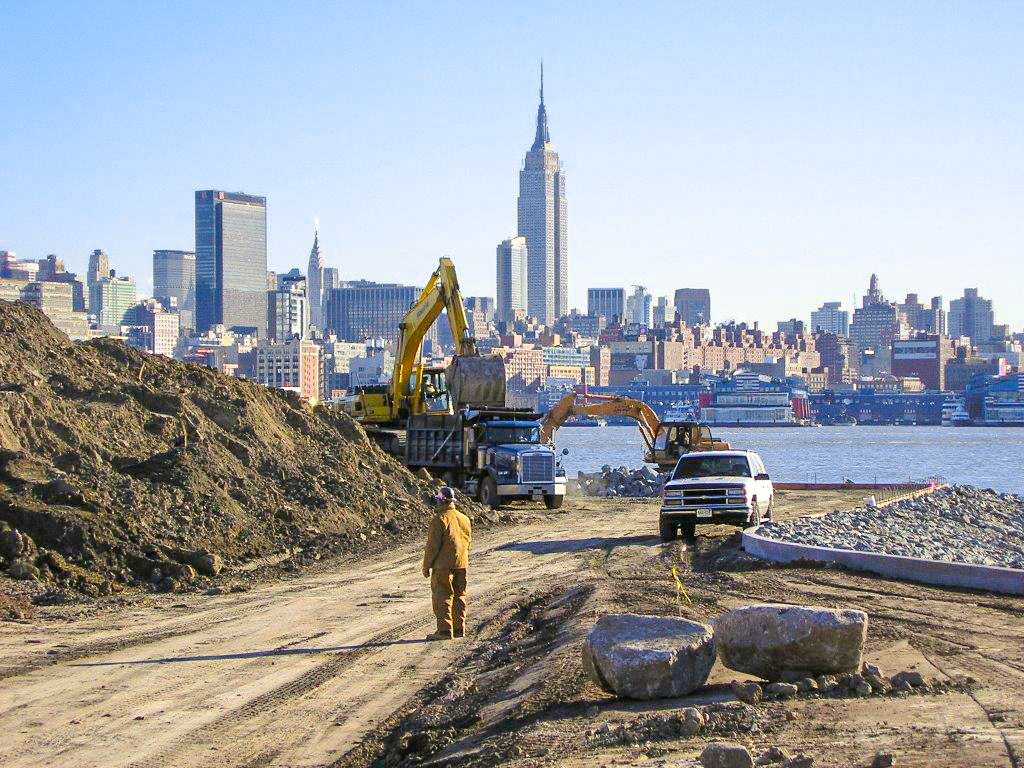 Excavators and trucks working on a waterfront construction site with a city skyline in the background.