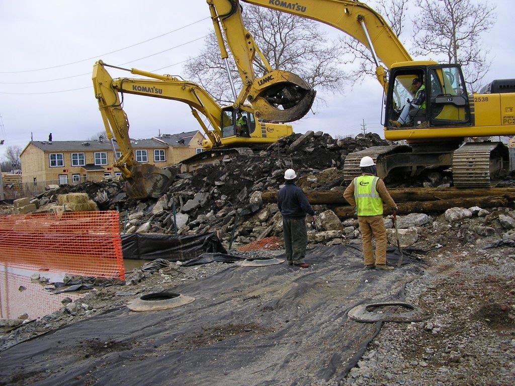 Excavators removing concrete and debris at a construction site while workers in hard hats supervise.