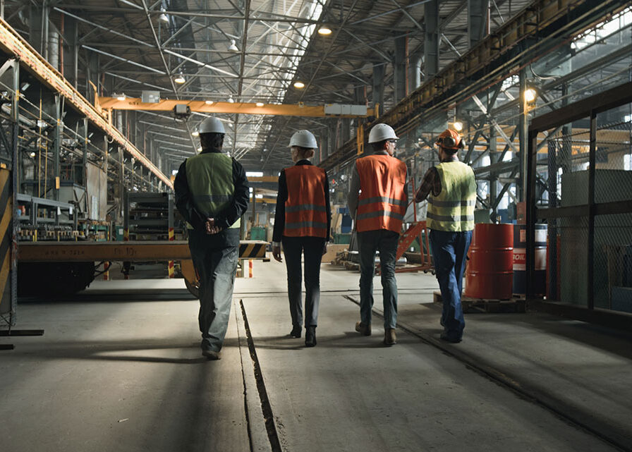 Industrial workers wearing hard hats and safety vests walking through a large manufacturing facility.
