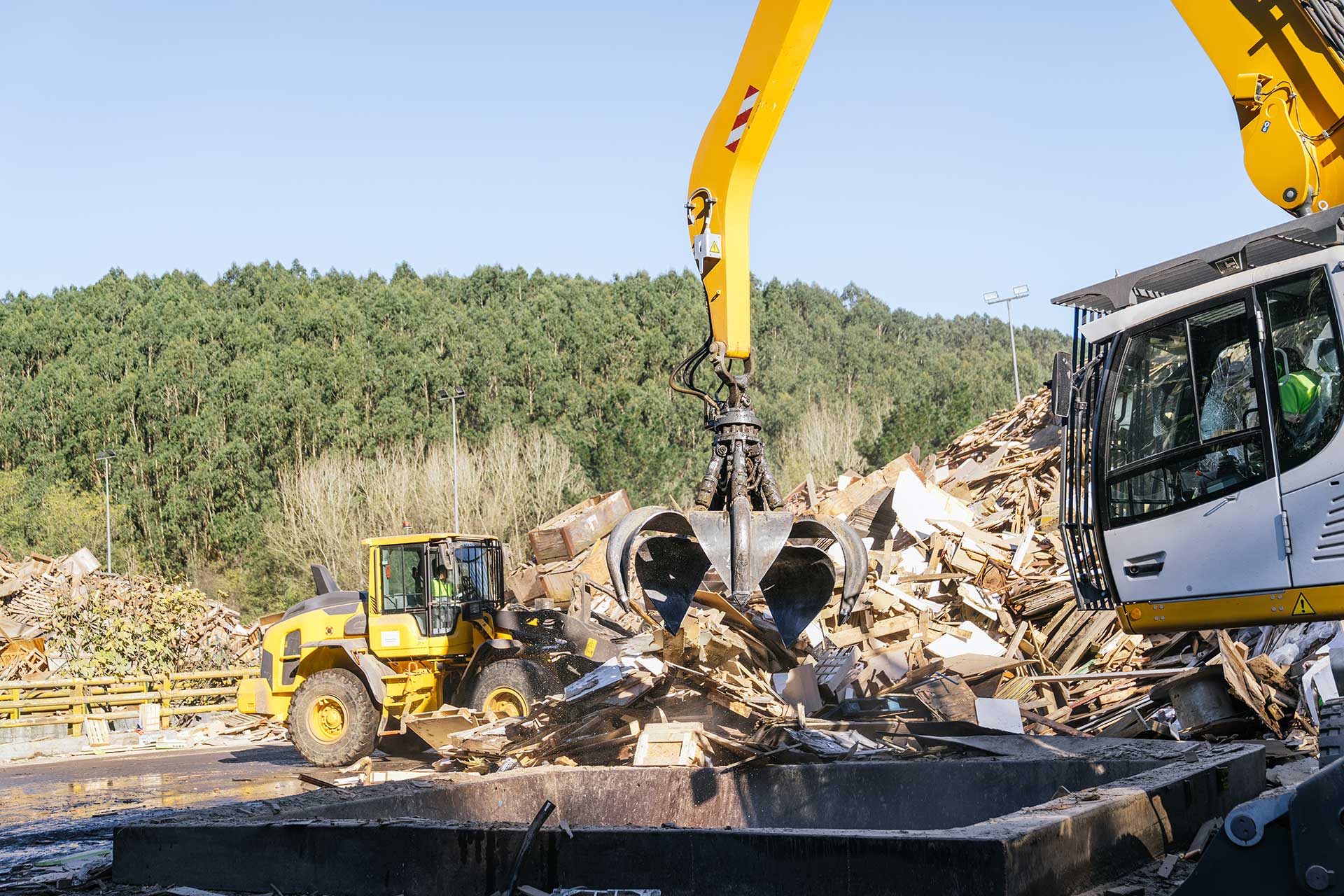 Excavator with grapple attachment sorting and loading wood debris at an industrial recycling facility.