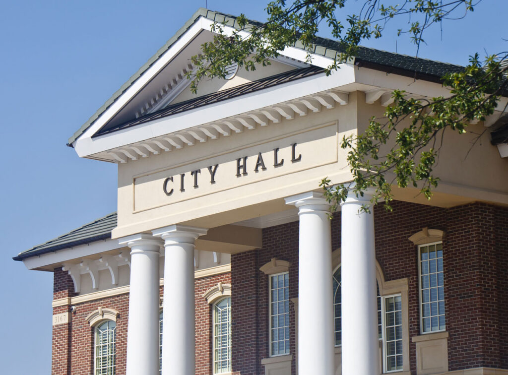 City Hall building with white columns, brick exterior, and classical architectural details under a blue sky.